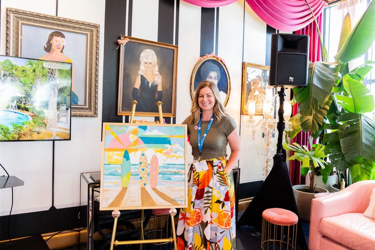 A woman stands beside a colorful beach-themed painting on an easel, surrounded by framed portraits and plants in a decorated indoor space.