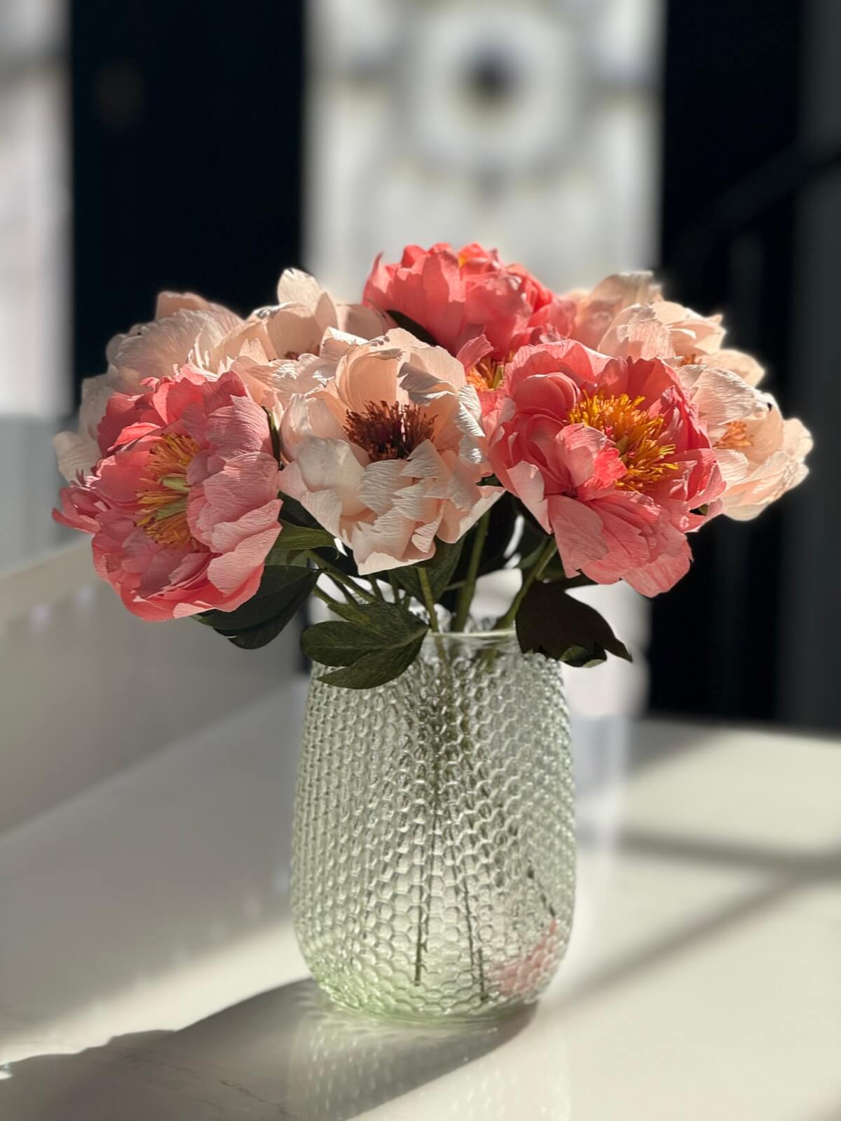 A clear textured glass vase holding pink and cream artificial flowers sits on a sunlit white surface—an elegant choice for Mother’s Day or thoughtful grad gifts.