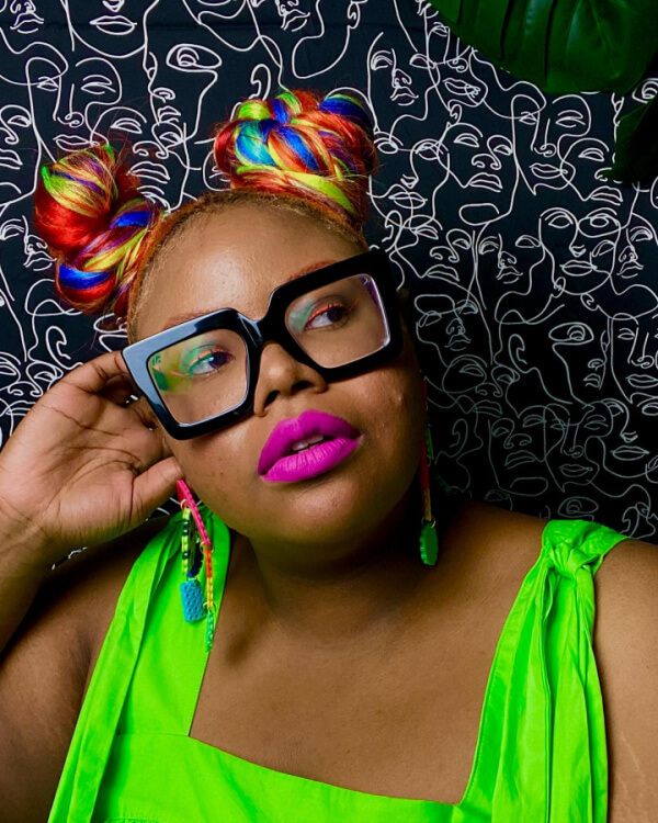 She's Behind BHAM's Most Colorful (and Secretly Nostalgic) Jewelry Line - 4 Aaliyah Taylor shows off rainbow hair buns, large black glasses, bright pink lipstick, a neon green top, and colorful earrings while posing in front of a black and white abstract line art background.