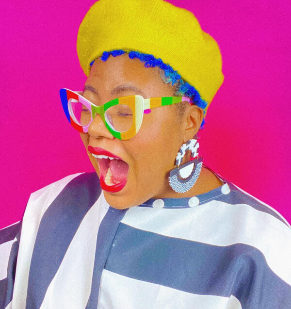 She's Behind BHAM's Most Colorful (and Secretly Nostalgic) Jewelry Line - 5 Aaliyah Taylor stands out in a yellow beret, colorful glasses, striped black and white clothing, and large earrings against a bright pink background.