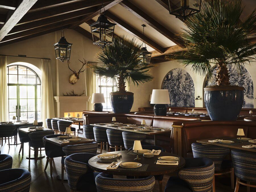 A cozy Houston dining area with round tables, striped chairs, large potted palm trees, arched windows, a fireplace, and decorative wall art under a beamed ceiling—perfect for fans of charming neighborhood restaurants.
