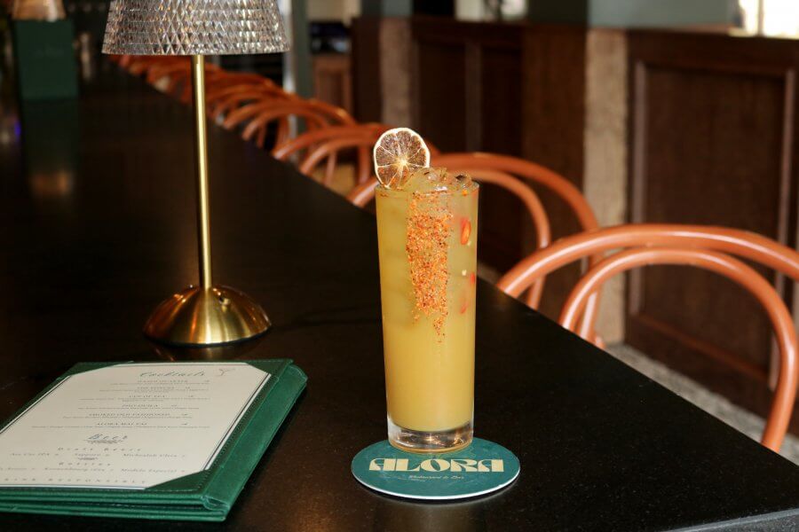 A tall yellow cocktail with ice and a dried citrus garnish sits on a coaster at a bar in one of the best restaurants Houston has to offer, with a menu and empty chairs visible in the background.