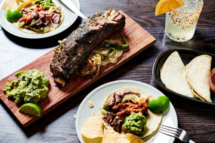 A wooden board with grilled ribs on sautéed onions, guacamole, and lime; nearby plates hold tacos, chips, and guacamole.