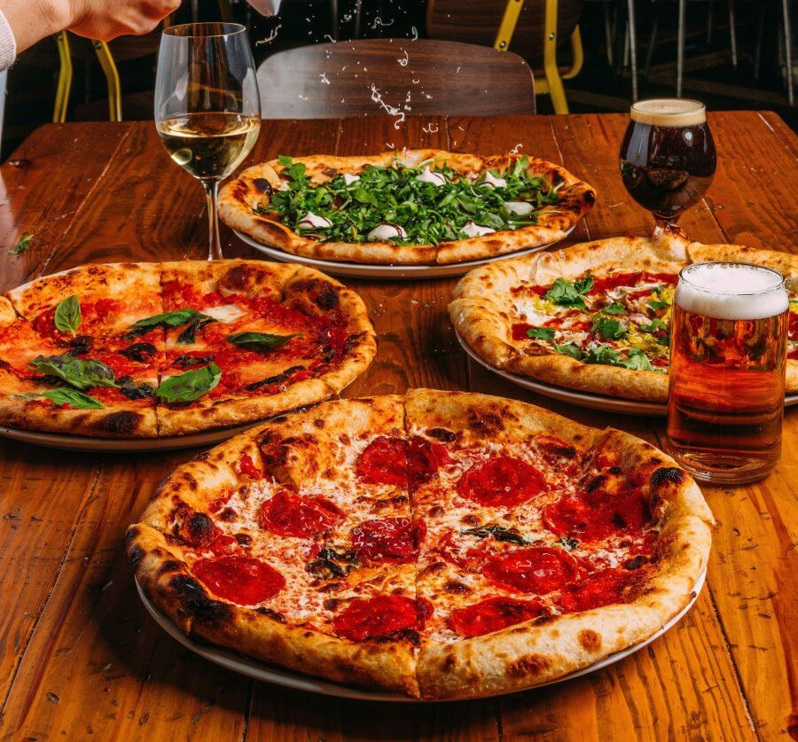 Four pizzas on a wooden table with three glasses of beer, a glass of white wine, and a hand sprinkling cheese onto an arugula pizza.
