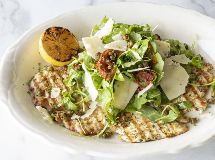 Grilled chicken breast topped with arugula, shaved parmesan, sun-dried tomatoes, capers, and a grilled lemon half on a white plate—a flavorful choice among standout Houston dining.