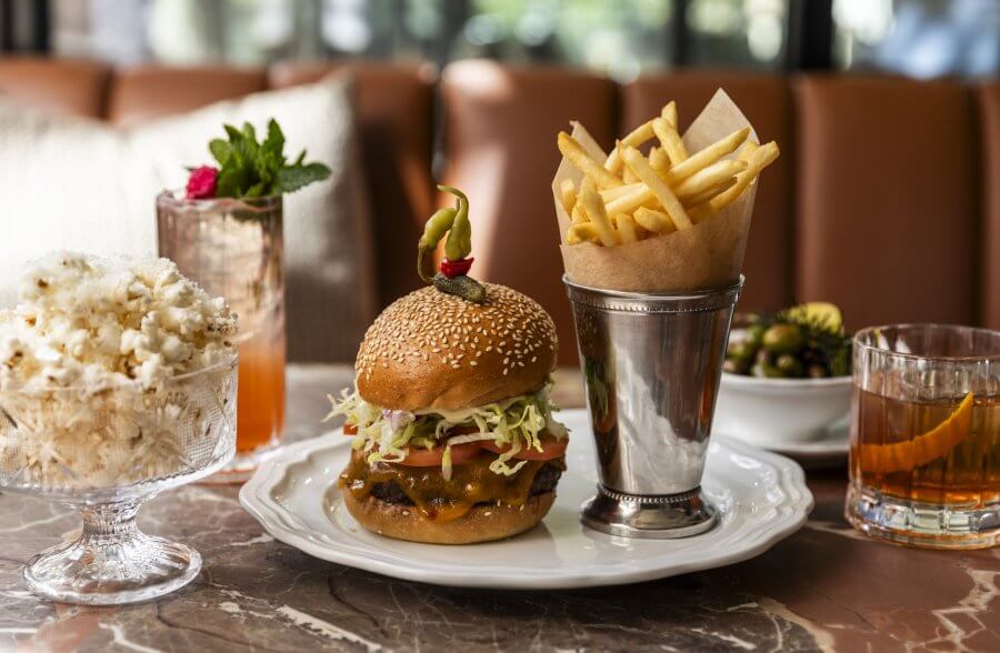 A plate with a cheeseburger topped with lettuce and tomato, a metal cup of French fries, a glass bowl of popcorn, and two cocktails on a marble table.