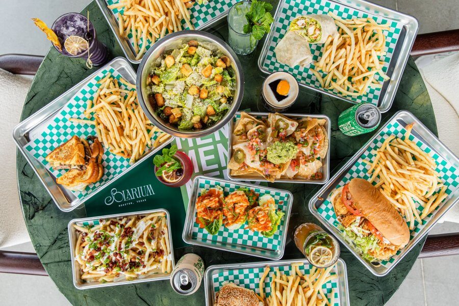 A table set with various trays of fries, sandwiches, tacos, a salad, drinks, and condiments on green checkered paper at a Houston neighborhood restaurant.
