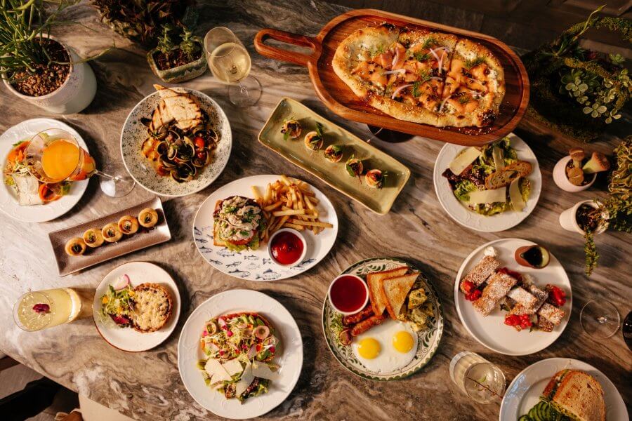 An overhead view of a marble table at one of Houston’s new neighborhood restaurants, set with plated dishes—flatbread, salads, sandwiches, fries, desserts, drinks—and surrounded by small potted plants.