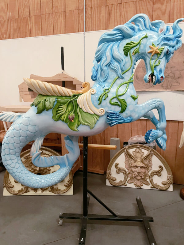 A painted carousel seahorse with a blue body, leafy green decor, golden wings, and floral details is displayed indoors on a metal stand at the Factory at Franklin's Carousel of Dreams.