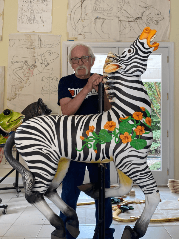 An older man stands behind a painted zebra carousel figure with orange flowers, inside the Factory at Franklin’s art-filled room, evoking the creative spirit of the Carousel of Dreams.