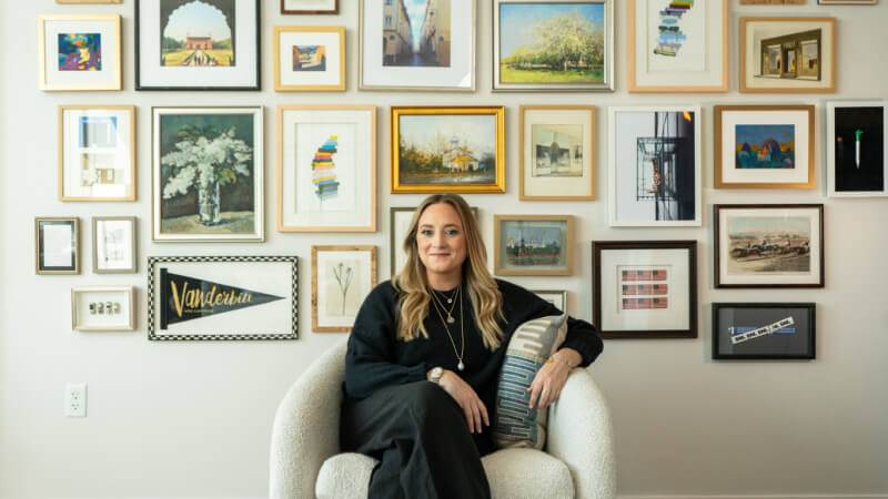 A woman sits in a white chair, holding a pillow, with a large gallery wall of framed artwork arranged behind her.