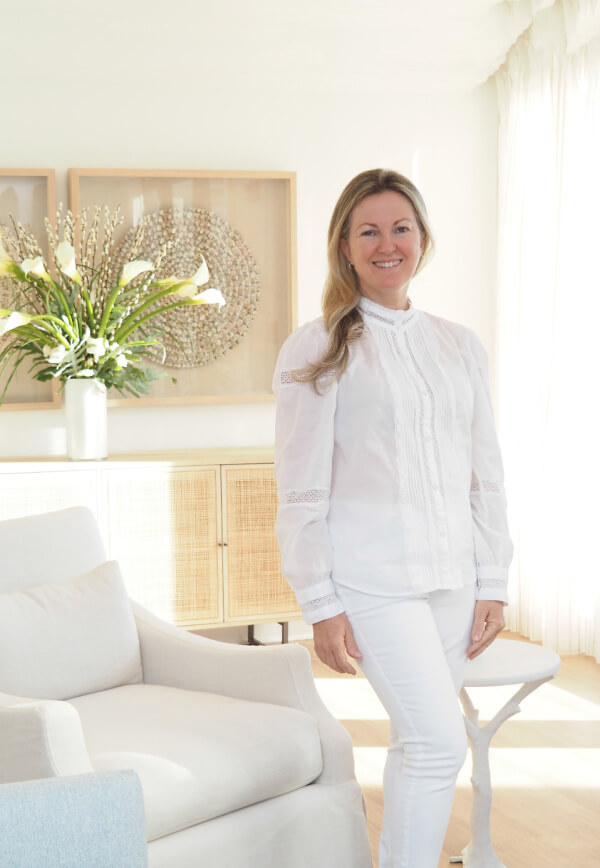 A woman dressed in a white blouse and white pants stands in a bright, modern living room with neutral decor, a vase of lilies on the sideboard, and her favorite Hollywood skincare products elegantly displayed nearby.