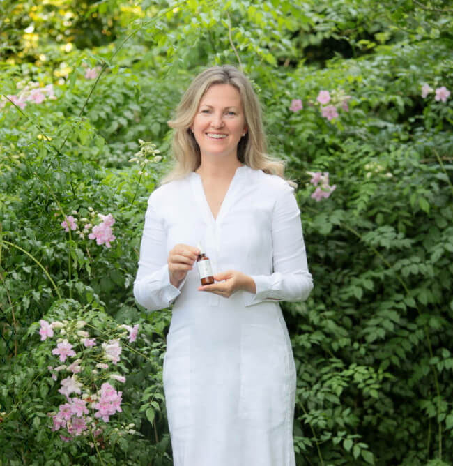 A woman in a white dress stands outdoors among green foliage and pink flowers, holding a small bottle from a skincare line and smiling at the camera.
