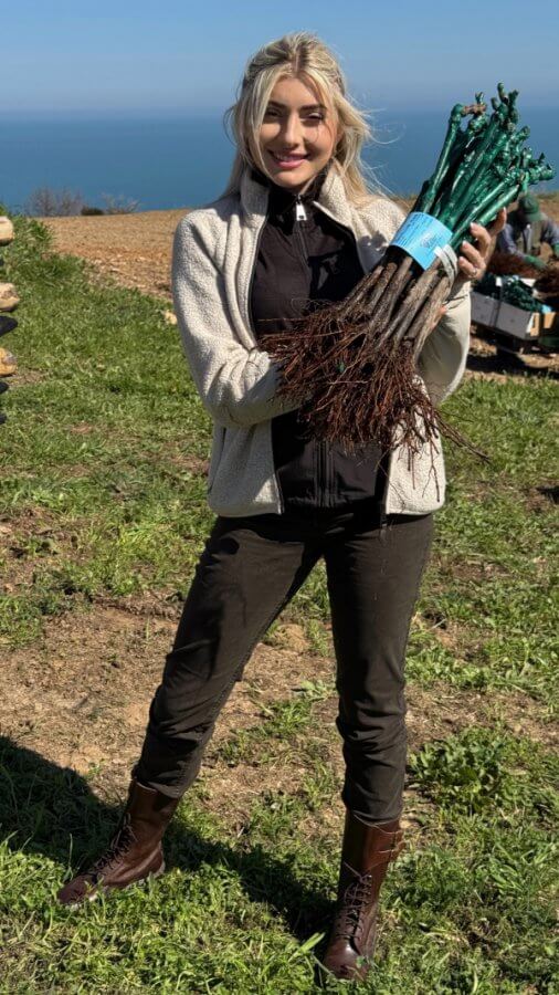 A person stands outdoors on grassy ground, holding a bundle of young plants with roots exposed. The ocean is visible in the background.