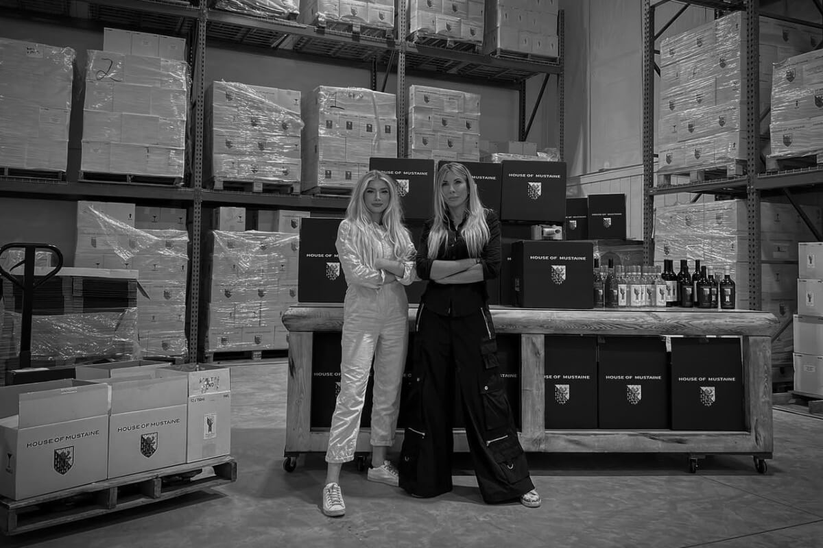 Pam Mustaine and Electra Mustaine stand side by side in a warehouse filled with stacked boxes and bottles on shelves, behind a table labeled "House of Mustaine."