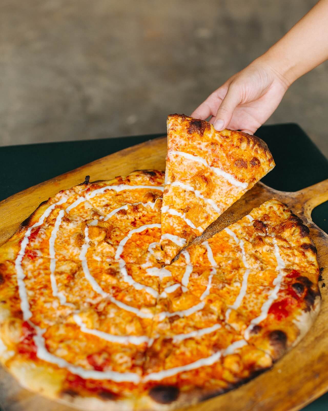 9 New Nashville Restaurants to Try This Month - 9 A hand lifts a slice of pizza with a drizzle of white sauce from a wooden pizza board, capturing the fresh flavors at Dukos.