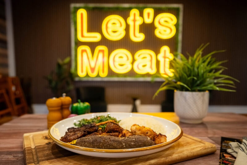 9 New Nashville Restaurants to Try This Month - 11 A plate of grilled meat and vegetables sits on a wooden table, with a "Let's Meat" neon sign and a potted plant in the background.