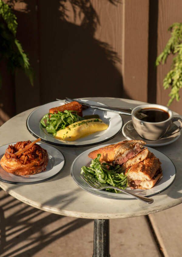 9 New Nashville Restaurants to Try This Month - 6 A round table set with three plates of food and a cup of black coffee; dishes include a pastry, a sandwich with salad, and a plate with greens, polenta, and a rectangular portion.