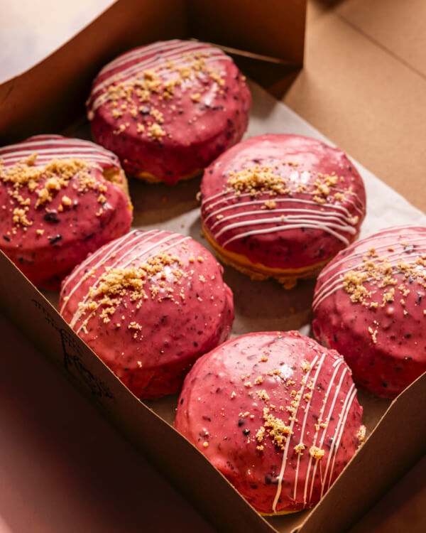 9 New Nashville Restaurants to Try This Month - 12 Six round pastries with pink glaze, white drizzle, and crumb topping arranged in a brown cardboard box—perfect for sampling creative flavors.