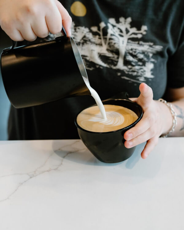 9 New Nashville Restaurants to Try This Month - 13 A person pours steamed milk into a cup of coffee, creating latte art with both hands holding the black cup and pitcher on a white countertop.