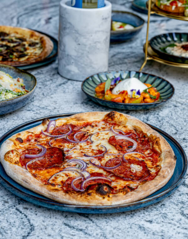 9 New Nashville Restaurants to Try This Month - 17 A cooked pizza topped with pepperoni and red onion is served on a blue plate, surrounded by various side dishes on a marble table—perfect for sampling at new Nashville restaurants opening in May 2025.