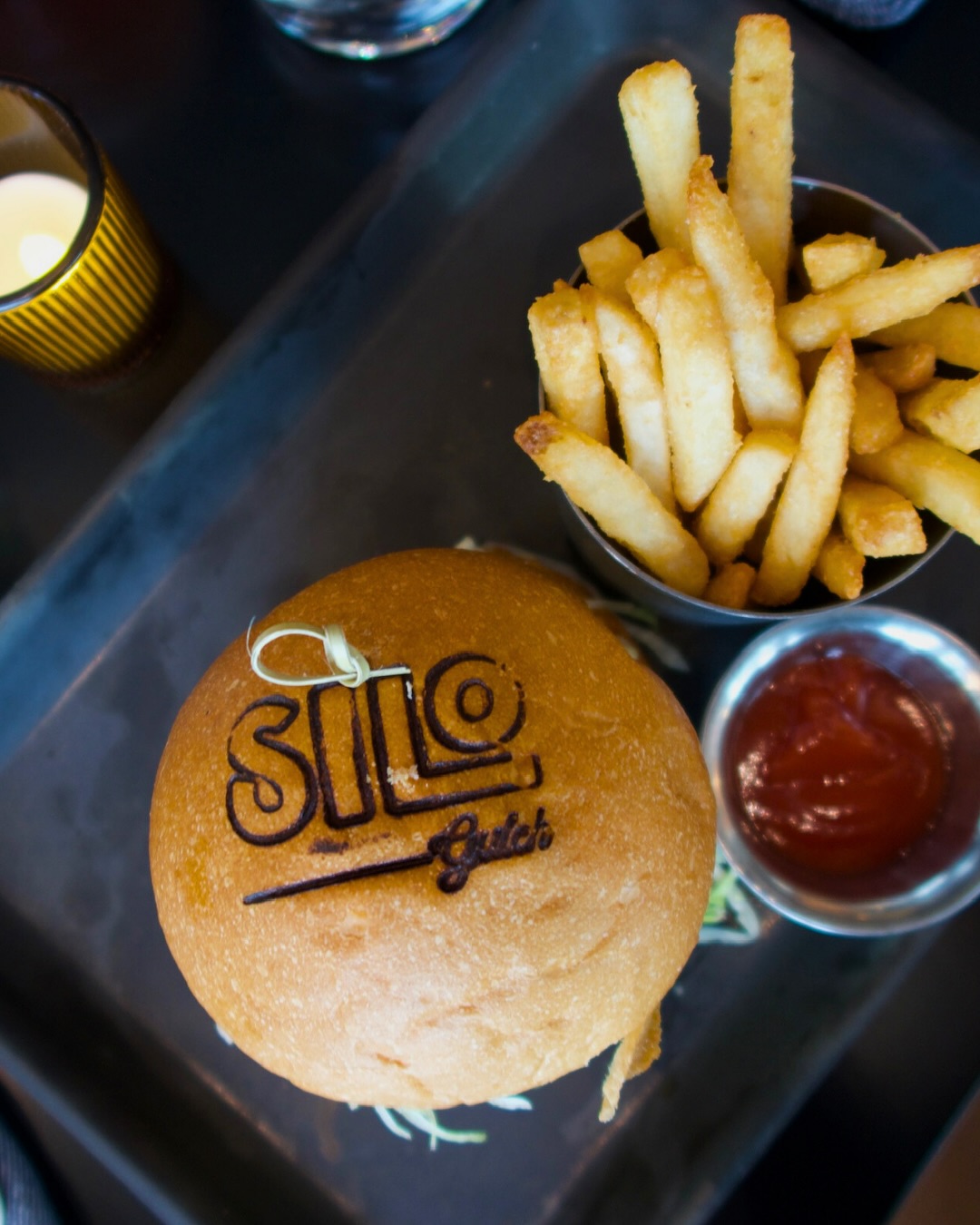 9 New Nashville Restaurants to Try This Month - 7 A hamburger with "Silo Guild" branded on the bun, served with fries and a metal dish of ketchup on a dark tray—an inviting bite among new Nashville restaurants.