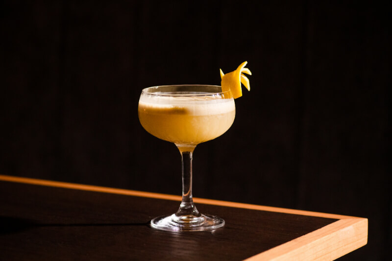 9 New Nashville Restaurants to Try This Month - 19 A yellow cocktail with a frothy top, served in a coupe glass and garnished with a lemon twist, sits on a dark wooden surface.