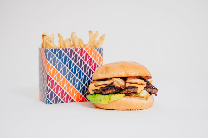 A cheeseburger with lettuce and bacon sits next to a container of French fries in a colorful box, set against a plain white background.
