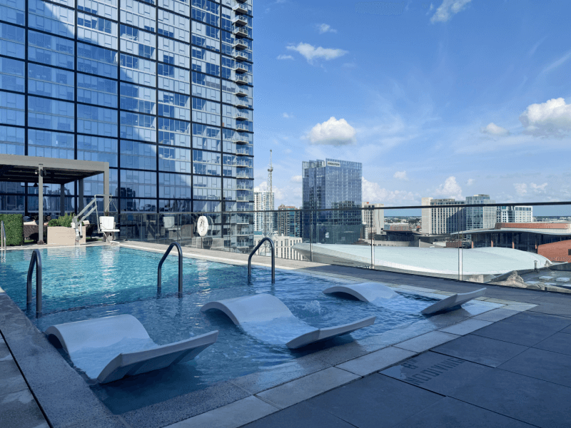 A rooftop hotel pool with three lounge chairs in the water, surrounded by a glass railing, overlooks city skyscrapers under a blue sky with scattered clouds.