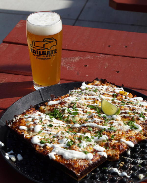 A rectangular pizza with white sauce drizzle and herbs sits on a black tray next to a pint of TailGate Brewery beer on an outdoor wooden table.