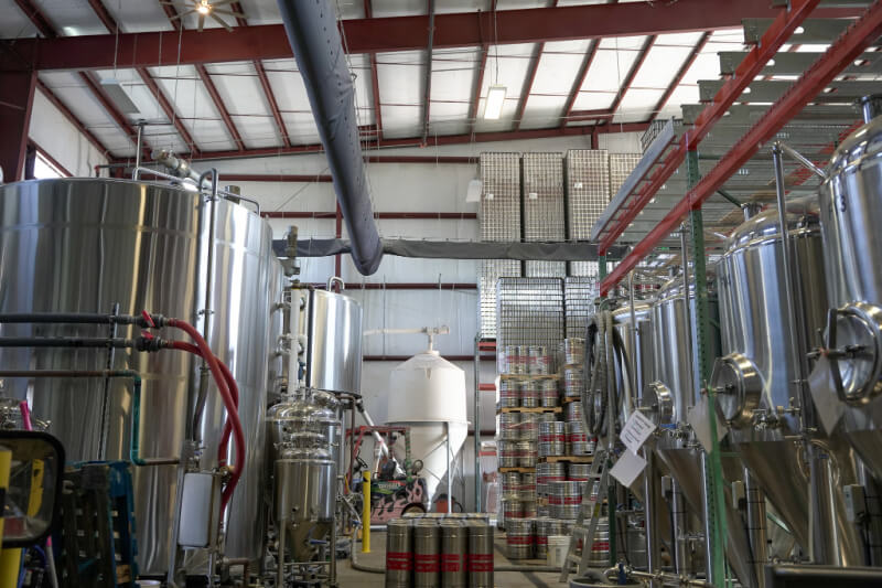 Interior of TailGate Brewery featuring large stainless steel tanks, stacked metal kegs, and rows of empty cans under a high, metal-beam ceiling.