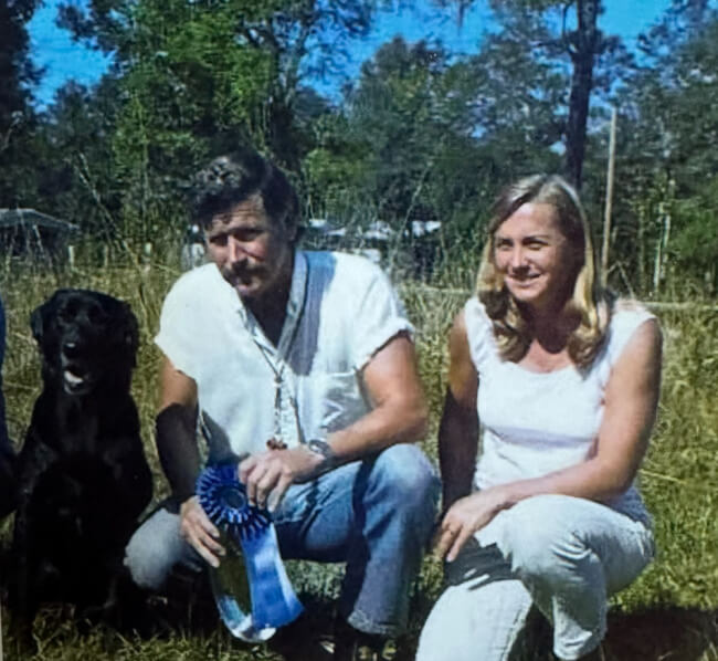 A man and a woman kneel in a grassy field with a black dog from Tealbrook Kennels; the man proudly holds a blue ribbon award.
