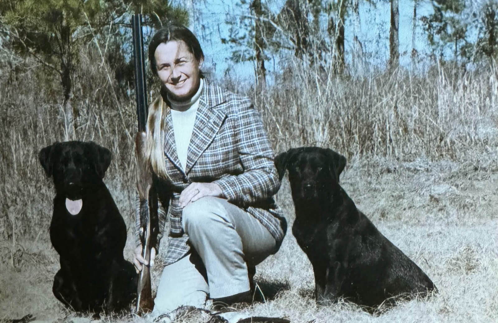 A person in a plaid jacket kneels outdoors holding a shotgun, with two black dogs. Trees and brush are visible in the background.