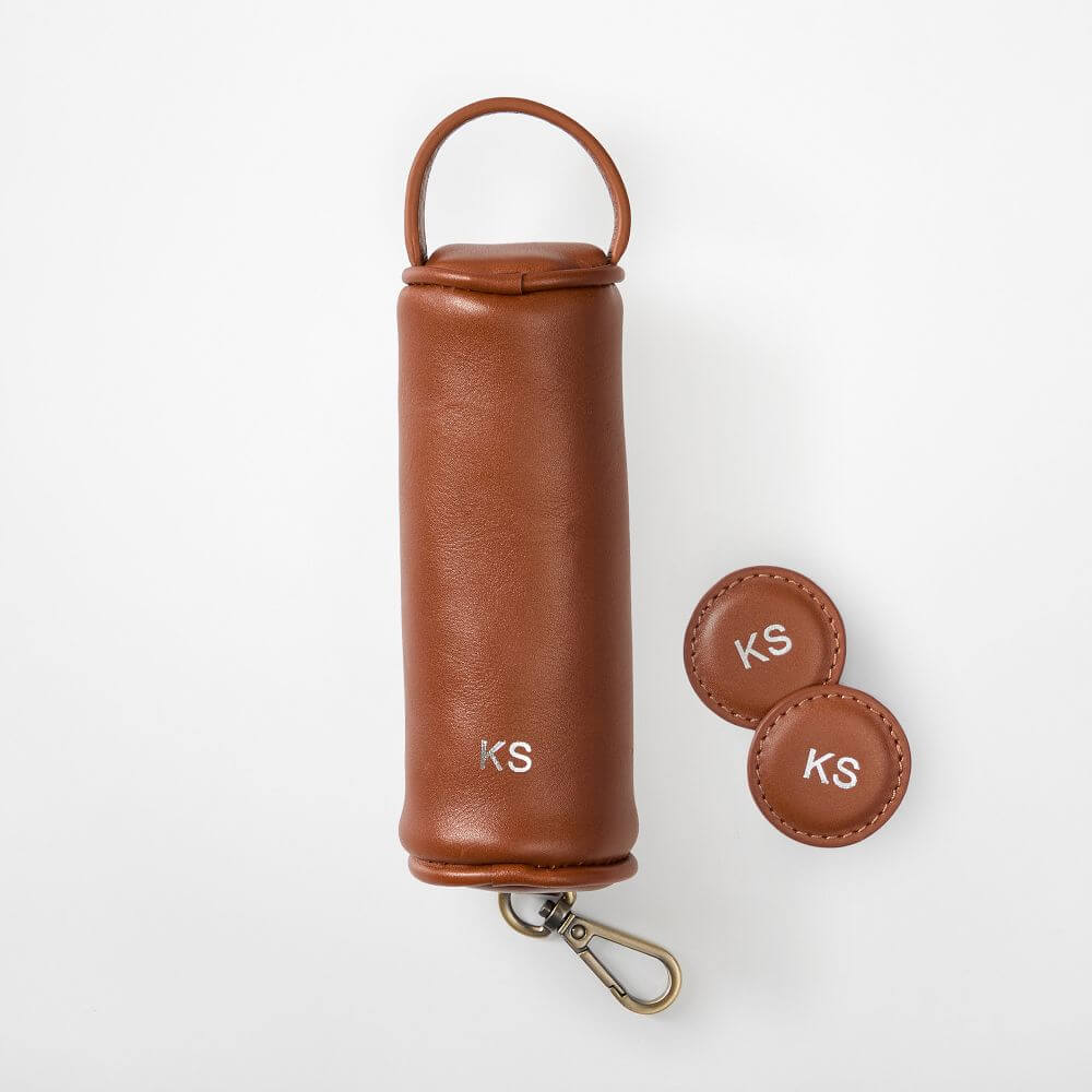 A brown leather cylindrical pouch with a handle and metal clasp, monogrammed “KS,” alongside two matching round leather keychains is a stylish pick for Father’s Day gifts or featured in any thoughtful gift guide.