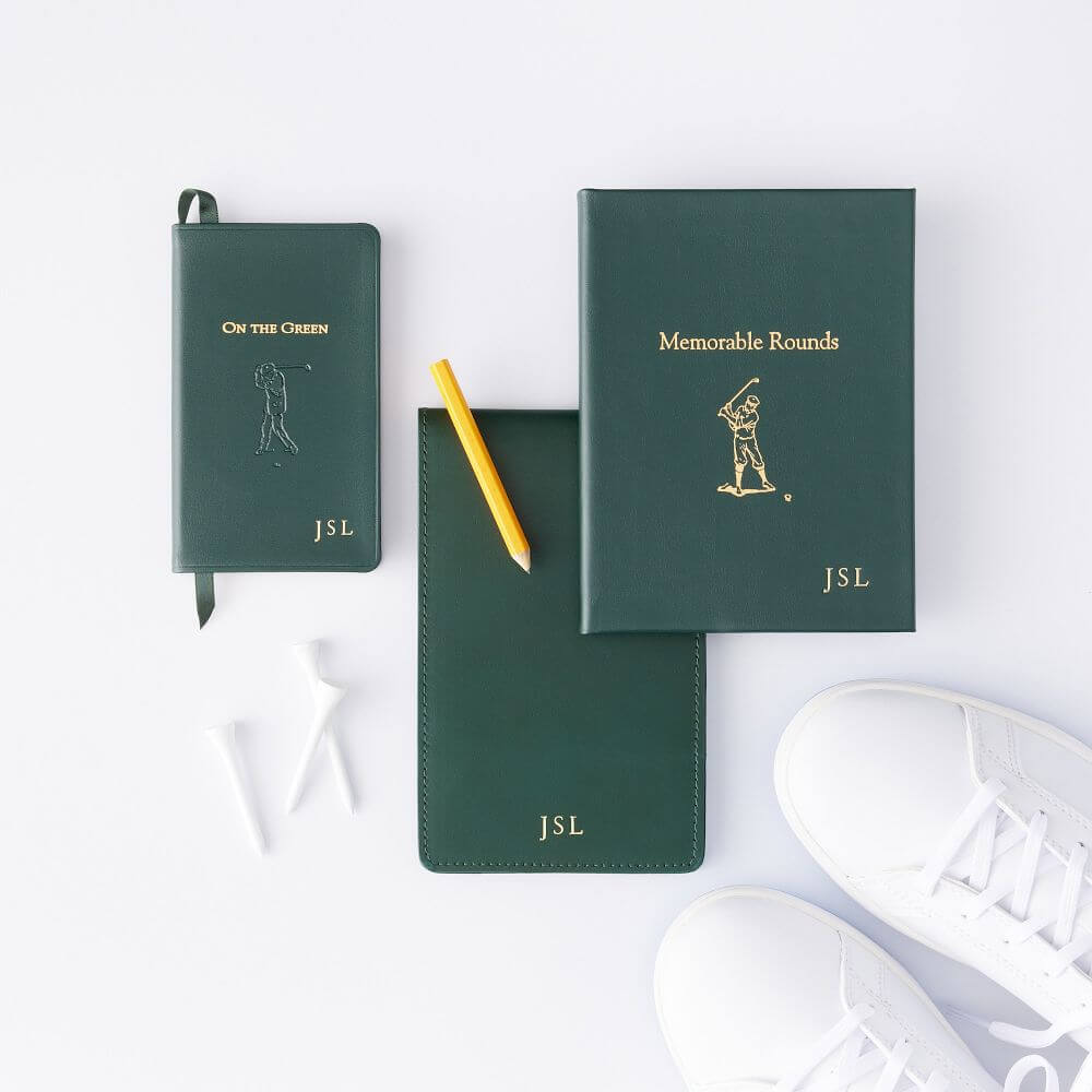 Three dark green golf-themed notebooks with gold lettering and "JSL" initials, a yellow pencil, white golf tees, and white sneakers are arranged on a white surface—ideal golf gifts for any enthusiast or as Father’s Day gifts.