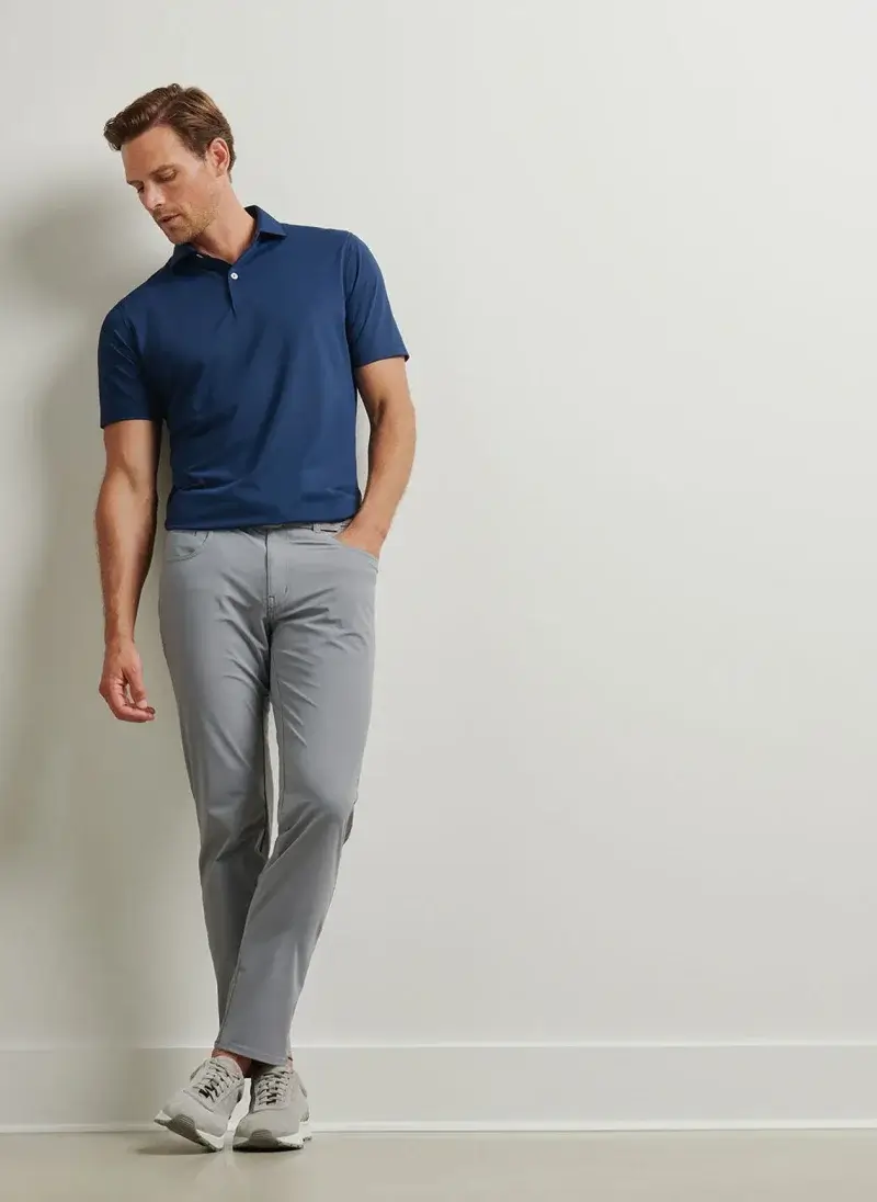 A man wearing a blue polo shirt, gray pants, and white sneakers stands against a plain white wall with one hand in his pocket—perfect inspiration for your Father’s Day Gift Guide or summer vacation style.
