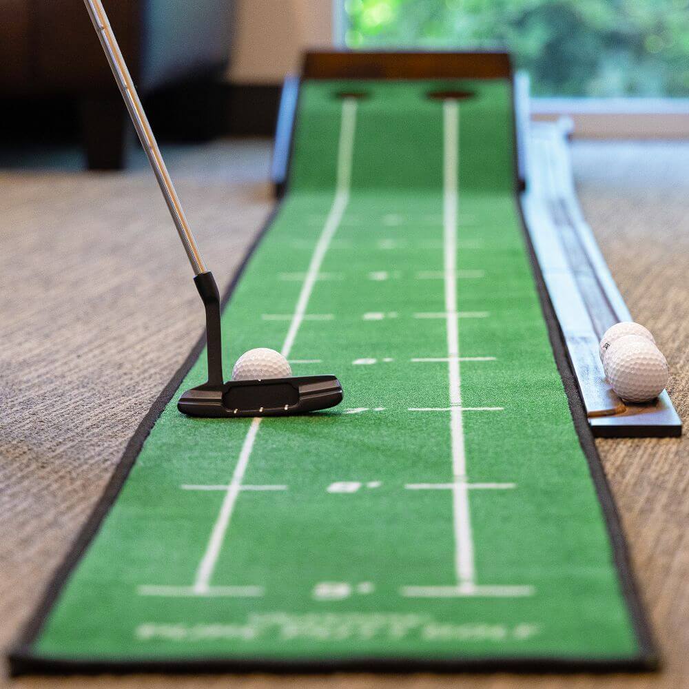 A person uses a putter to hit a golf ball on a green putting mat indoors, with two more golf balls beside the mat—an ideal pick for Father’s Day Gifts for the golf-loving dad.