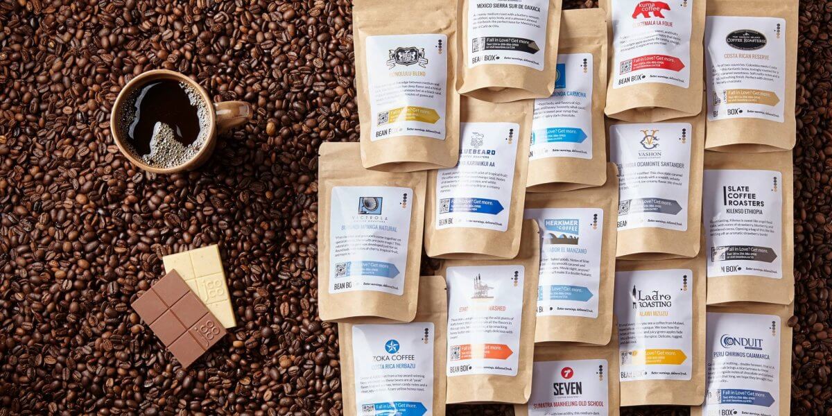 A cup of black coffee with a chocolate bar sits on coffee beans beside an array of assorted packaged coffee samples—perfect for Father's Day gifts or adding to your favorite subscription boxes.