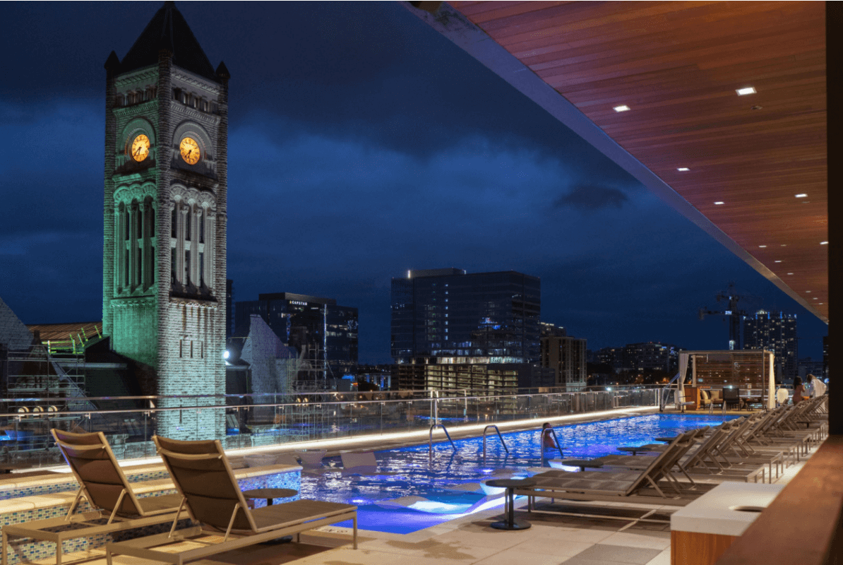 A rooftop pool with lounge chairs overlooks a cityscape at night during CMA Fest, featuring a clock tower with glowing faces and modern buildings in the background.