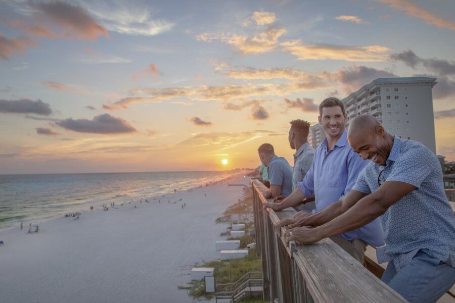 5 Reasons South Walton is Becoming the Premier Destination for Corporate Events and Meetings - 2 Four men stand and lean on a boardwalk railing at sunset, overlooking a sandy beach and ocean with a hotel building in the background—capturing the relaxed vibe of an event in South Walton.