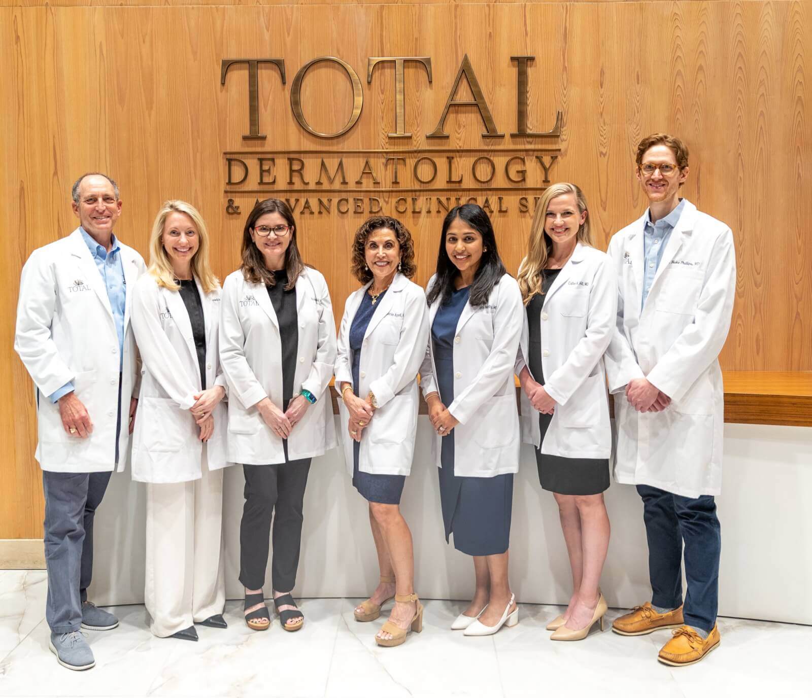 Seven medical professionals in white lab coats stand in a row, smiling, in front of a wall sign that reads "Total Dermatology & Advanced Clinical," reflecting the team’s dedication to expert dermatological care.