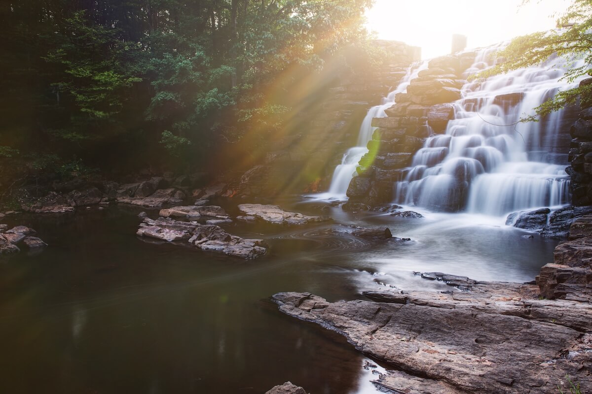 Water cascades down a rocky waterfall into a calm pool, surrounded by trees with sunlight streaming through the foliage—a serene scene reminiscent of hidden gems near classic SEC Towns.