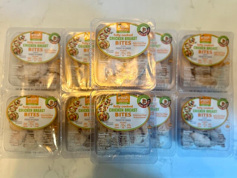 Ten packs of Fresh brand fully cooked chicken breast bites are arranged on a white surface. Each pack displays nutritional information and the Protein Picks at Costco product label.