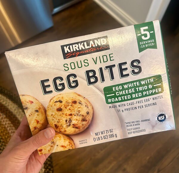 A hand holds a box of Kirkland Signature Sous Vide Egg Bites, featured as one of the Protein Picks at Costco. The egg white bites with cheese trio and roasted red pepper offer 5 servings and 11g protein per serving.