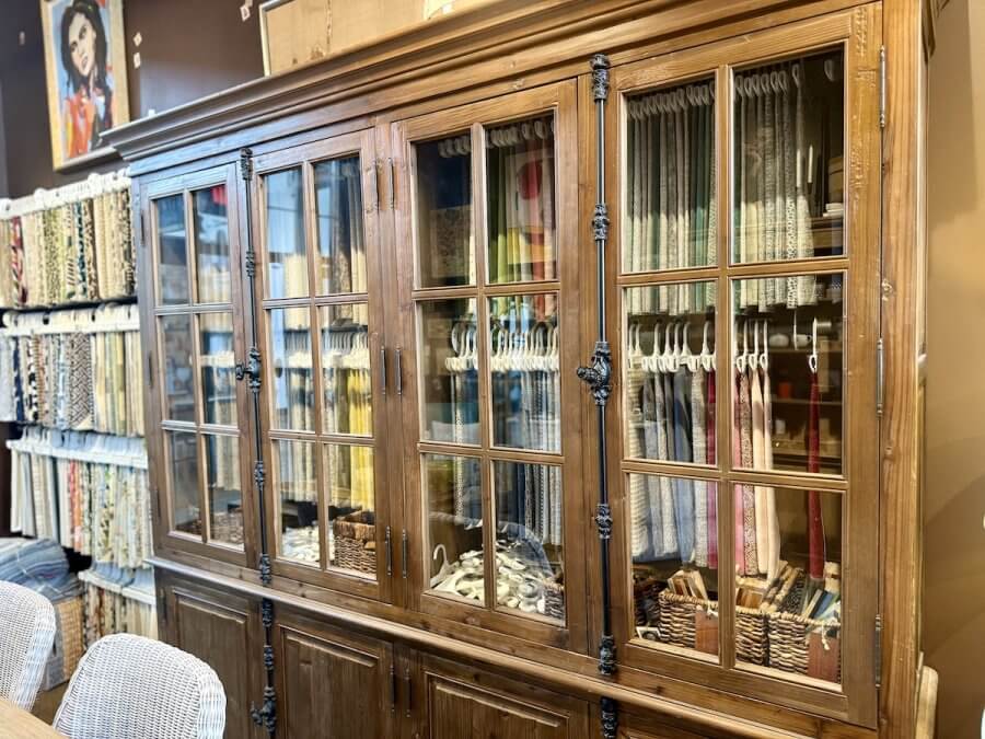 A wooden display cabinet with glass doors holds baskets and fabric samples, adding personality to the space. More fabric swatches hang on racks nearby in this well-lit room at a Nashville Home Store with warm brown walls.