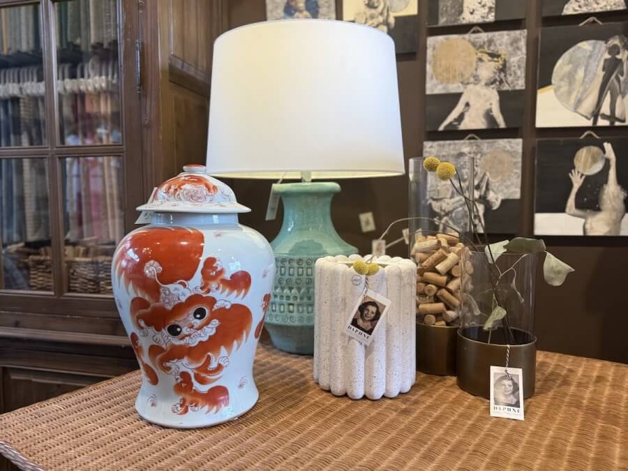 A decorative table from a Nashville home store features a red and white porcelain jar, green lamp, white textured vase, glass jar with dried flowers, and framed art on a brown wall—perfect for elevating your home decor.