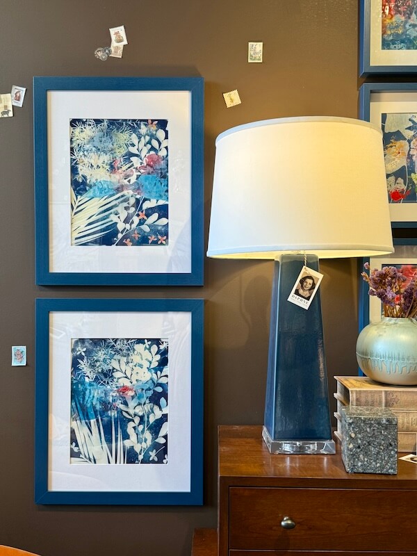 Framed botanical prints hang on a brown wall beside a blue lamp with a white shade, dried flowers, stacked books, and a decorative box—perfect inspiration for home decor from your favorite Nashville home store.