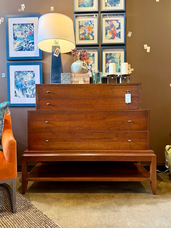 A wooden dresser with staggered drawers is displayed under a table lamp, in front of a wall with framed artwork and surrounded by decorative items, adding personality to this Nashville home store’s unique home decor collection.