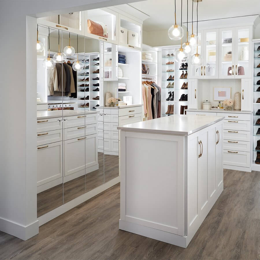 A spacious walk-in closet with white cabinetry, a central island, hanging clothes, shelves with shoes and bags, mirrored doors, and pendant lights—perfect for those seeking luxury closet design inspiration.