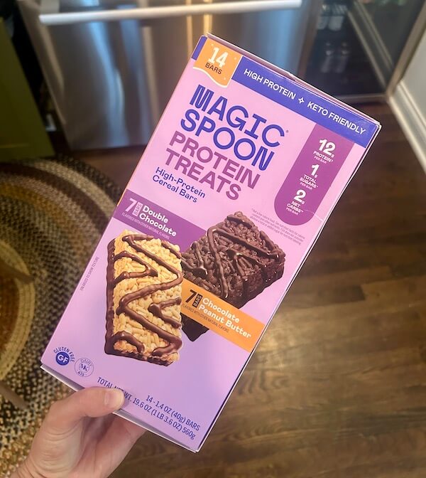 A hand holds a box of Magic Spoon Protein Treats, featuring 14 high-protein cereal bars in Double Chocolate and Chocolate Peanut Butter flavors—one of the top Protein Picks at Costco.